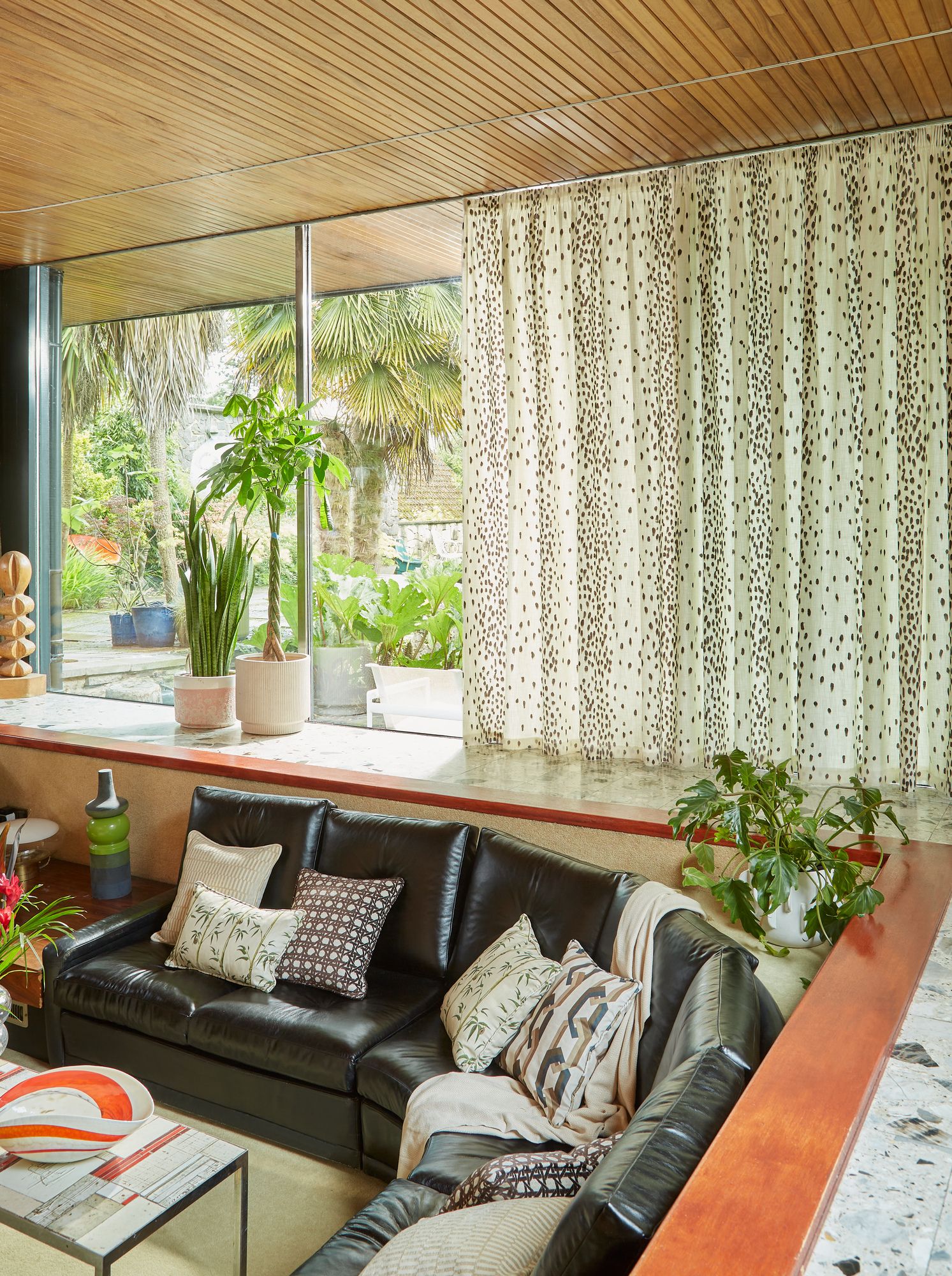A sunken living room with a curved, black leather sectional and patterned cushions, in front of large windows and spotted curtains. Indoor plants enhance the bright, mid-century modern space.