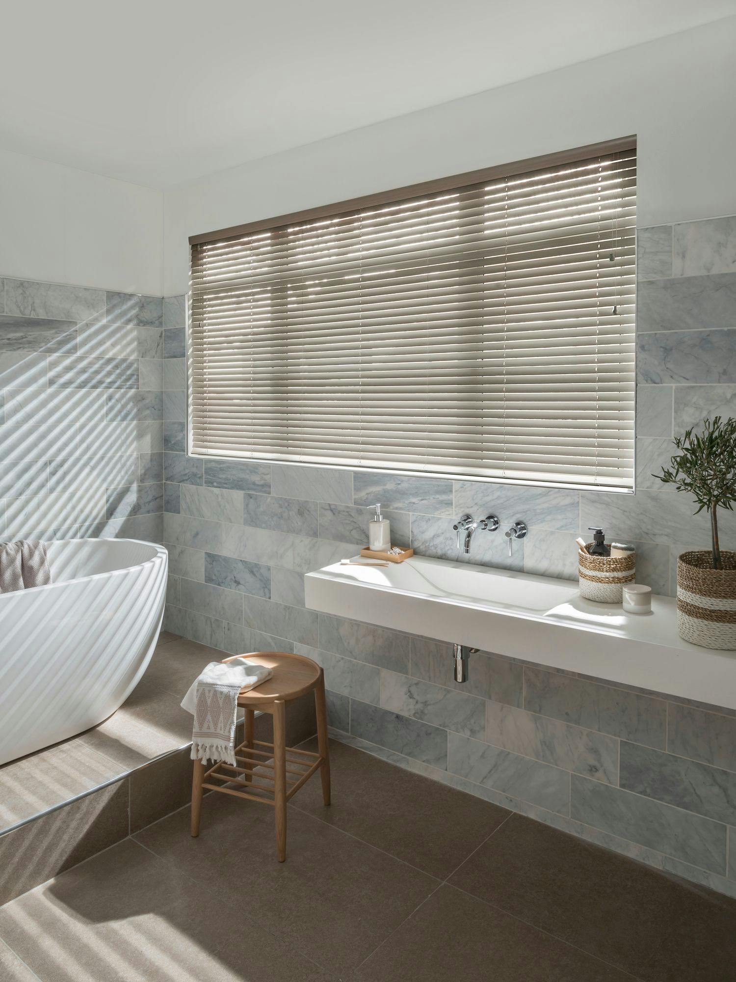 Dusk-grey faux-wood venetian blinds on a bathroom window above a white tub, slats tilted down for privacy.