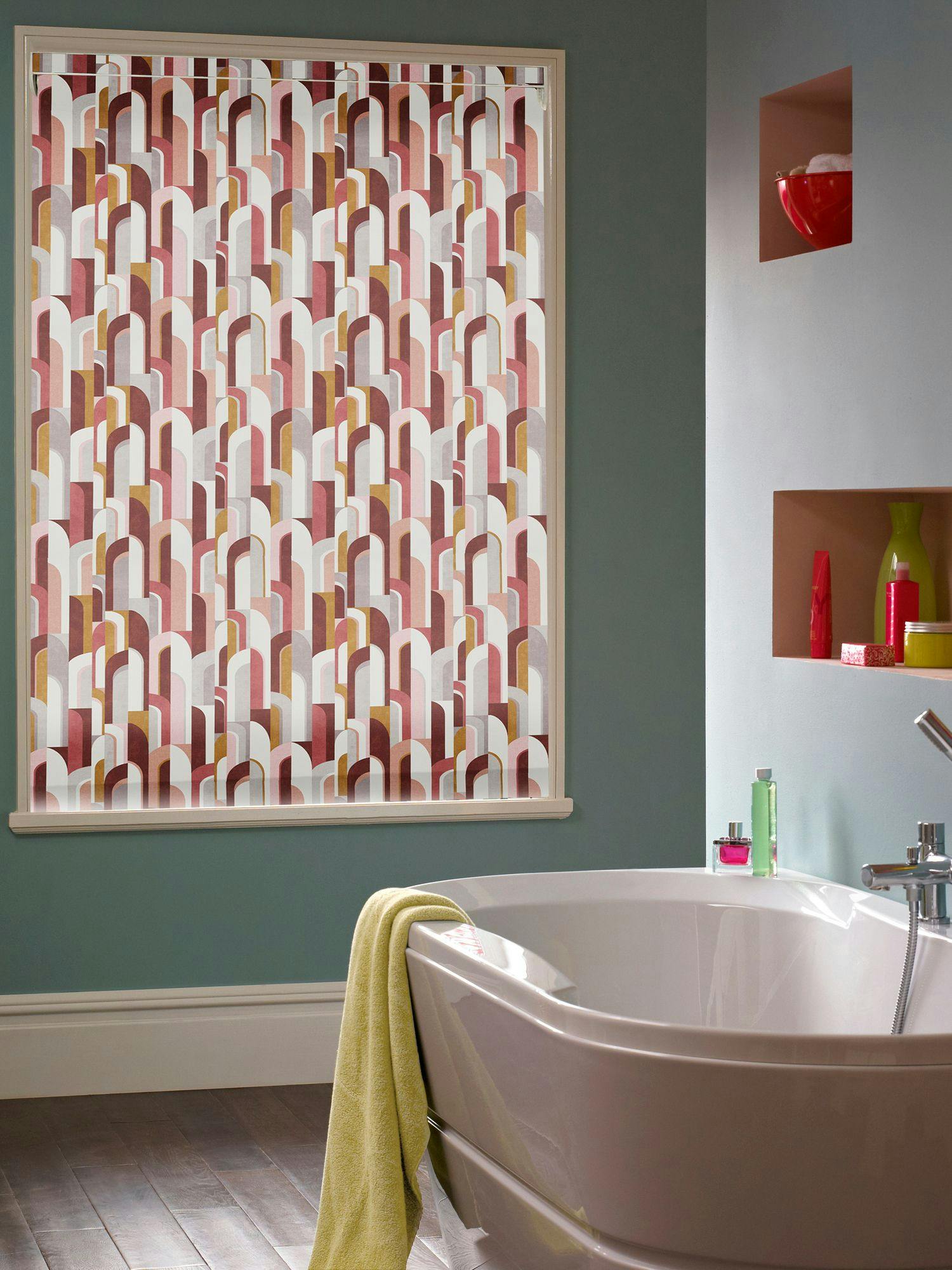 Retro roller blind with pink, mustard and terracotta Bauhaus‑style arches on a bathroom window behind a freestanding tub.