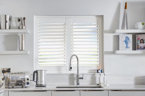 White kitchen with small window dressed in white vinyl shutters, closed with louvres open, behind a swan neck silver tap, pale grey base units with white and grey specked worktop and splashback, shelving to both sides with cookery books, and kitchen utensils, toaster and kettles next to sink.