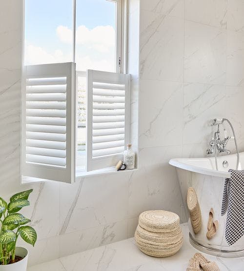 Bright bathroom with white café-style vinyl shutters, marble walls, a freestanding tub, woven stool, and green plant for a fresh, modern look.
