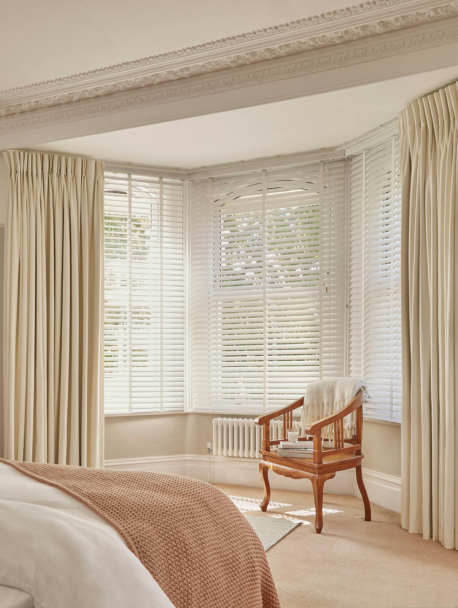 Ivory bedroom with decorative cornice and deep bay window dressed in white wooden venetian blinds, fully lowered and tilted, and ivory pleat curtains, fully open, wooden chair in recess with cream fringed throw, bed with cream bedding and light brown waffle throw.