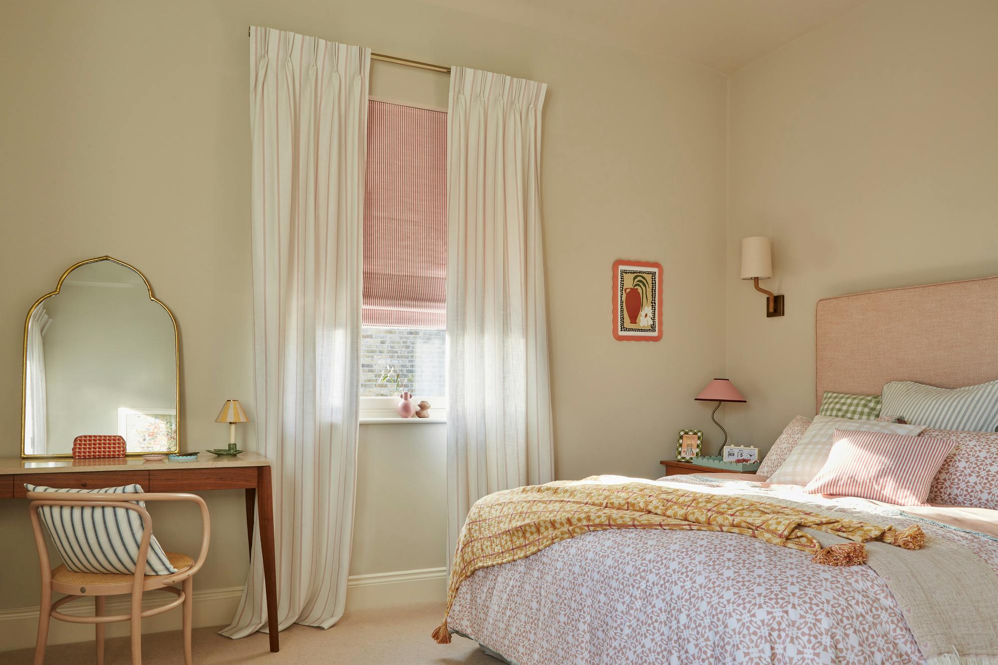 Bedroom with cream walls, bed with dusky pink headboard, bedding in white and pink patterned cover with cushions with stripes and checks, dressing table and shaped mirror and window dressed with pale red and white striped Roman blind with curtains in white with pale red pinstripe on a rod mounted higher above the window.