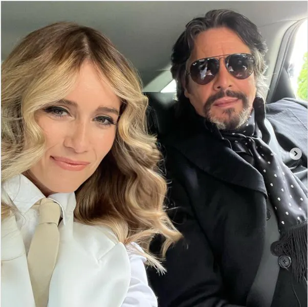 Laurence Llewelyn-Bowen wearing black jacket, black shirt and black silk scarf whith white dots and Whinnie Williams wearing white jacket white silk shirt and taupe tie sit together in the back seat of a car.