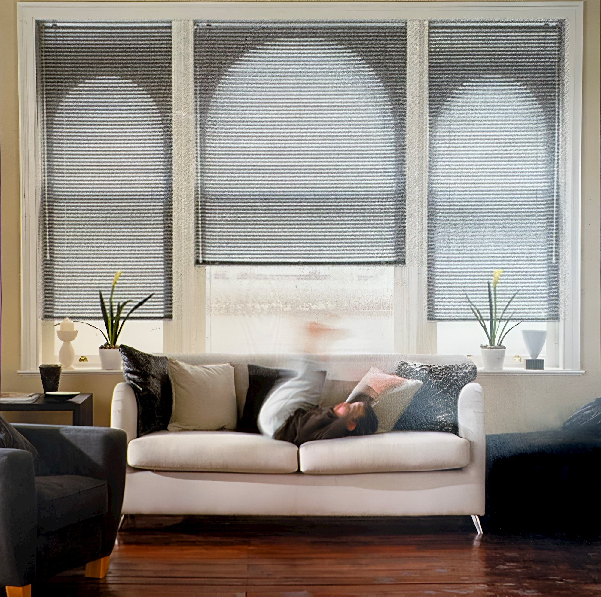 Close in on cream living room with wide window dressed in dark grey metal venetian blinds, centre blind lowered halfway, right and left blinds lowered three quarters, all tilted, cream modern sofa in front with cream, and dark grey cushions, a child rolls on the sofa, flowering plants on windowsill in white pots, wooden floor. 