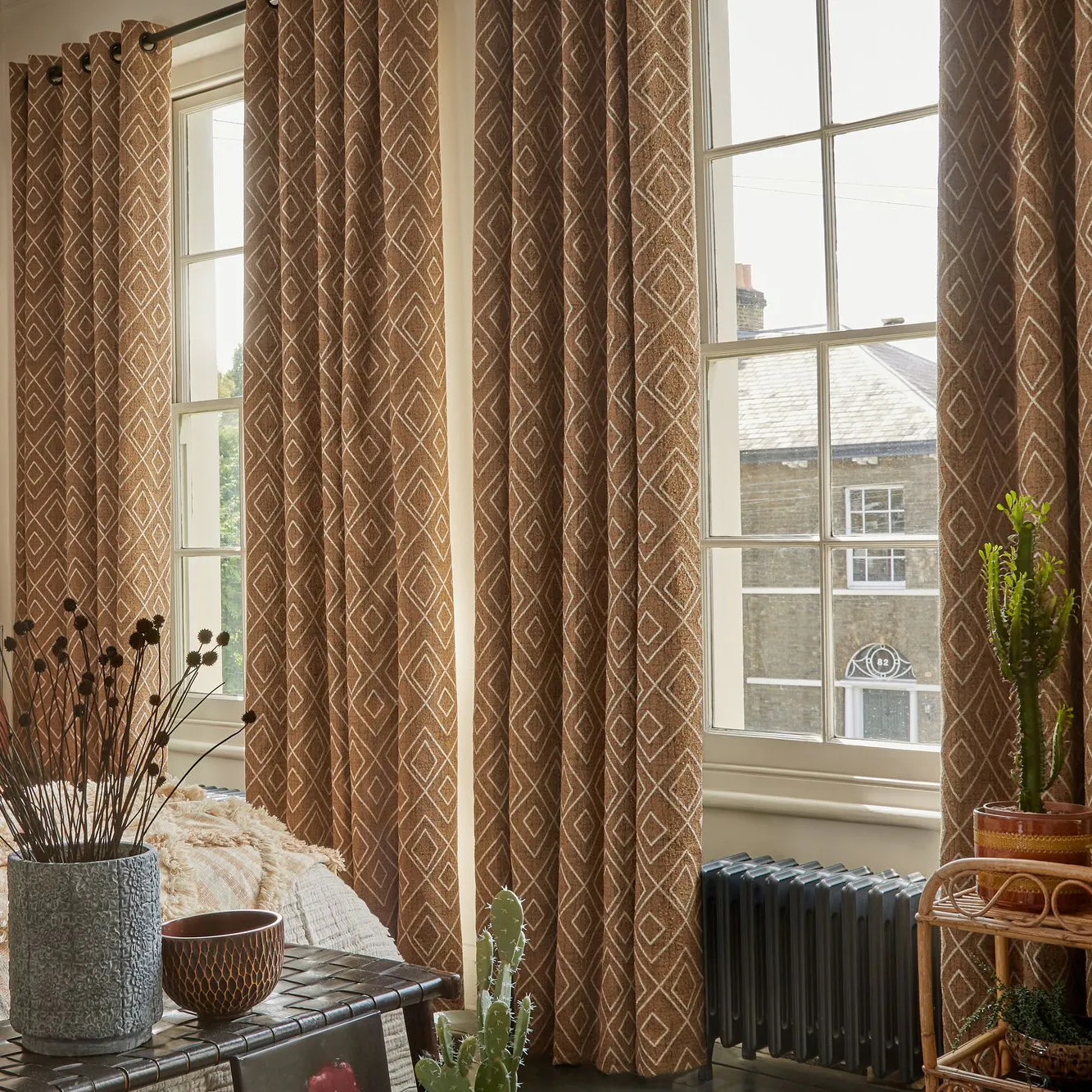 Taupe bedroom with two adjacent tall windows dressed with mid cinnamon curtains with a white diamond pattern, open, bed with turmeric cover and textured throw at foor in pale beige, cushions on bed in creams, charcoal and one matching curtains, pampas by bed in base, wood and weave bench at bed end with grey vase of dried dark flowers, cactus on books on floor.