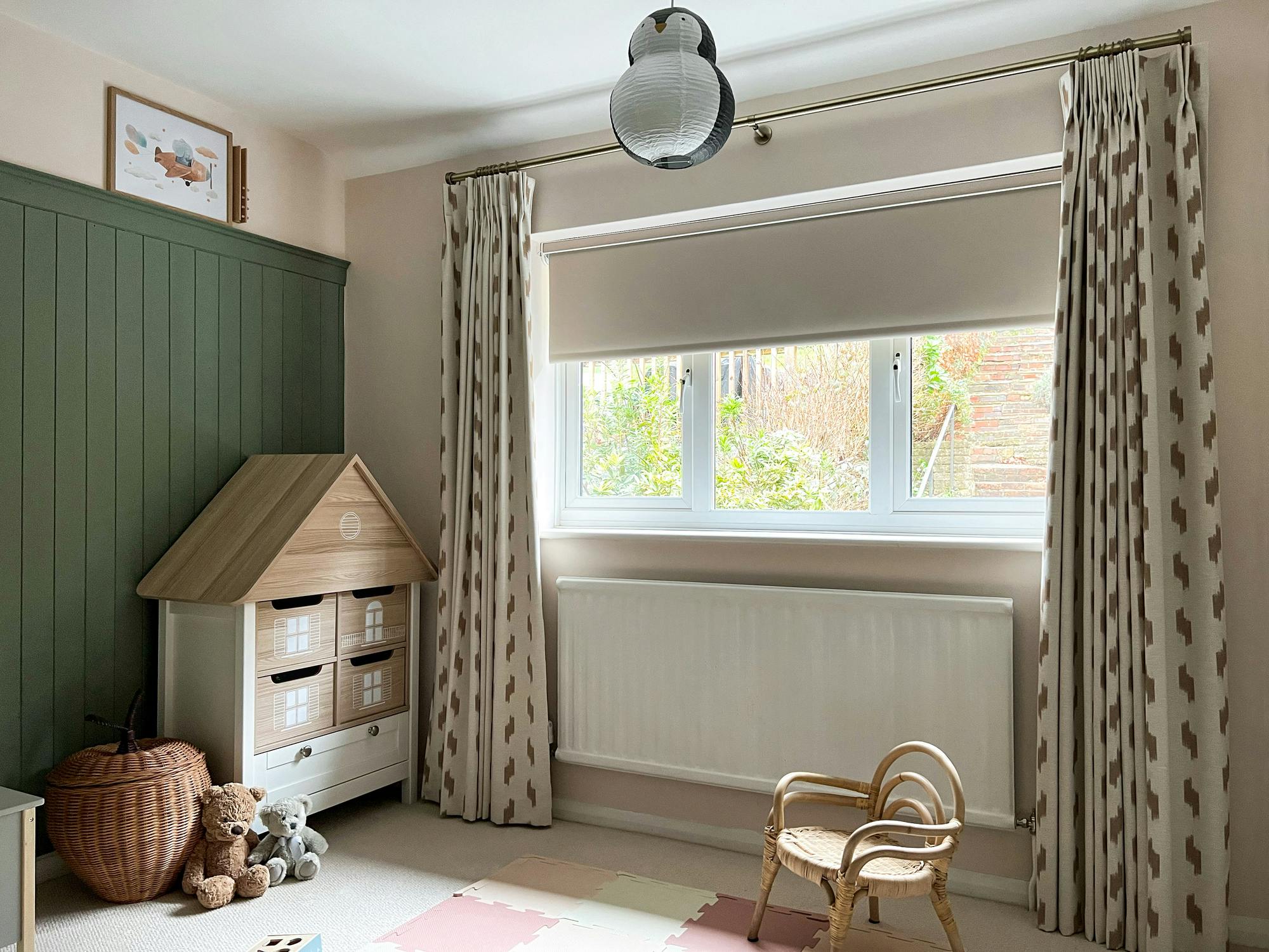 A wooden dollhouse shelf stands in a child's room with teddy bears on the floor, patterned curtains beside a window, and a small wicker chair, creating a cozy play area.