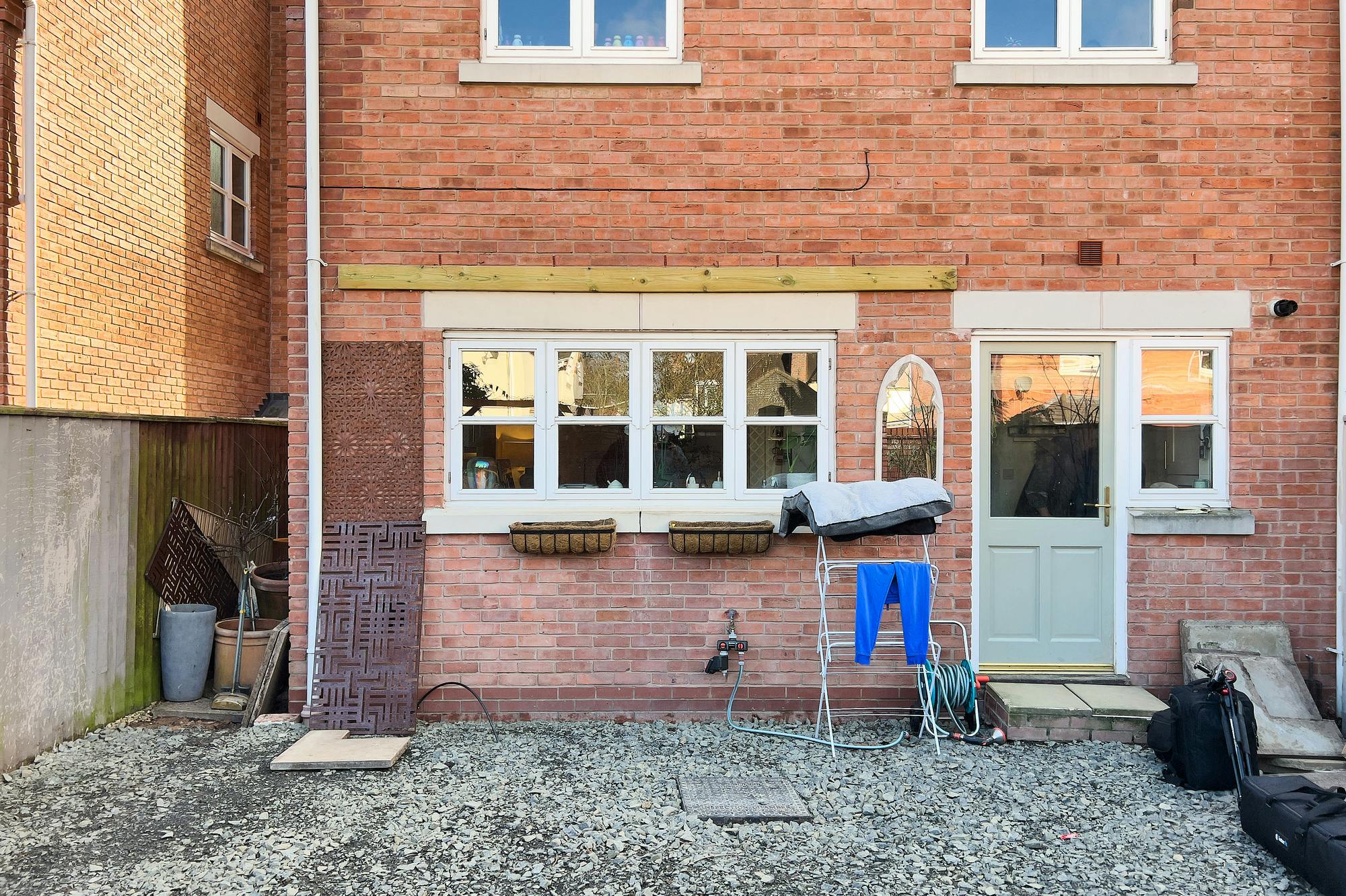 A brick house exterior with white-framed windows, a back door, and a gravel-covered yard, featuring a drying rack, garden hose, and some stored items.