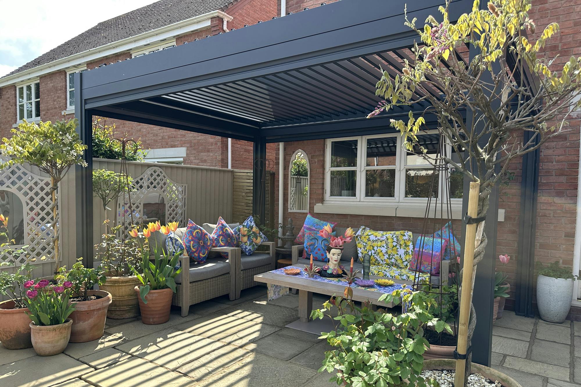 A stylish patio with a freestanding pergola, colorful cushions on outdoor seating, vibrant potted plants, and a decorative table in front of a brick house.