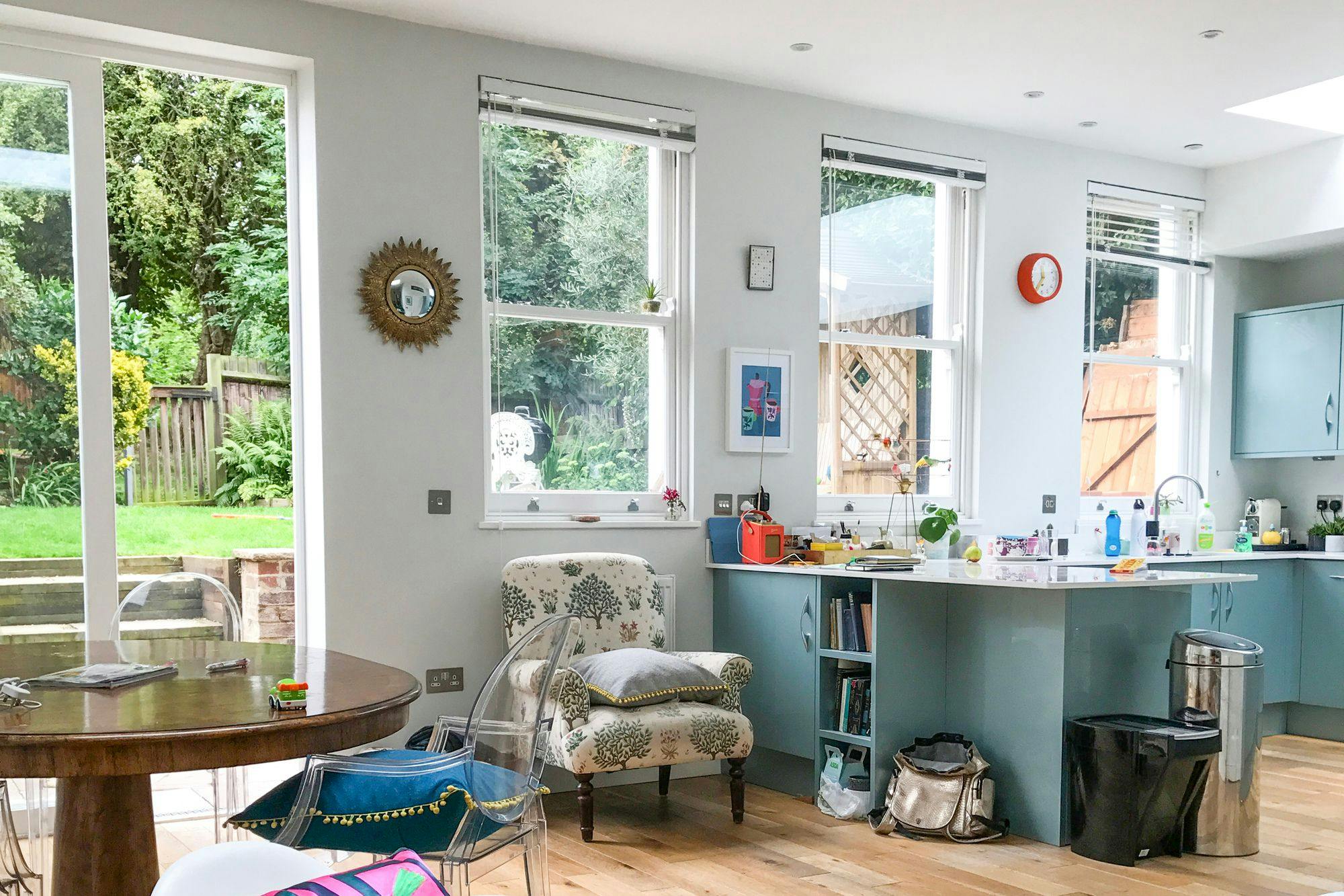 Bright kitchen with blue cabinets, wooden floors, and large bare windows letting in natural light, overlooking a garden.