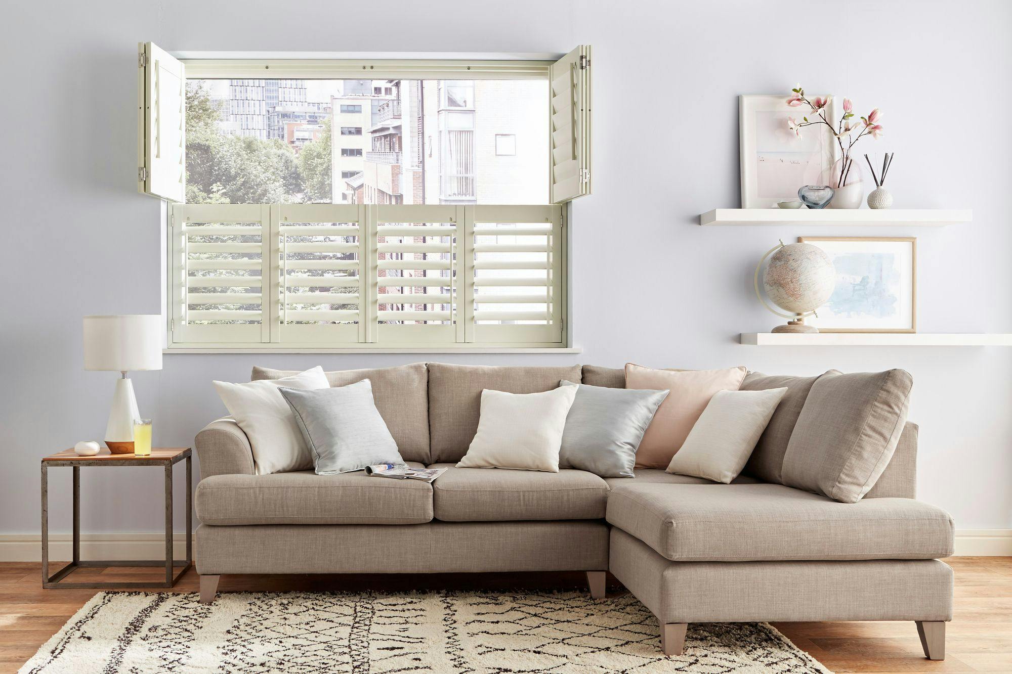 Neutral-toned living room with beige sectional sofa, white tier on tier shutters, decorative pillows, and floating shelves with art, flowers, and a globe.