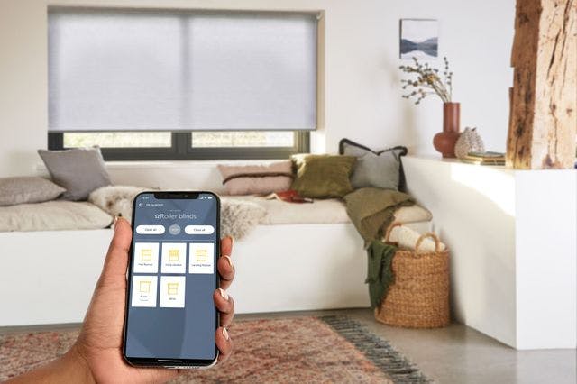 A hand holding a mobile device with Somfy app displayed on the screen in front of a window with light grey roller blinds on it.