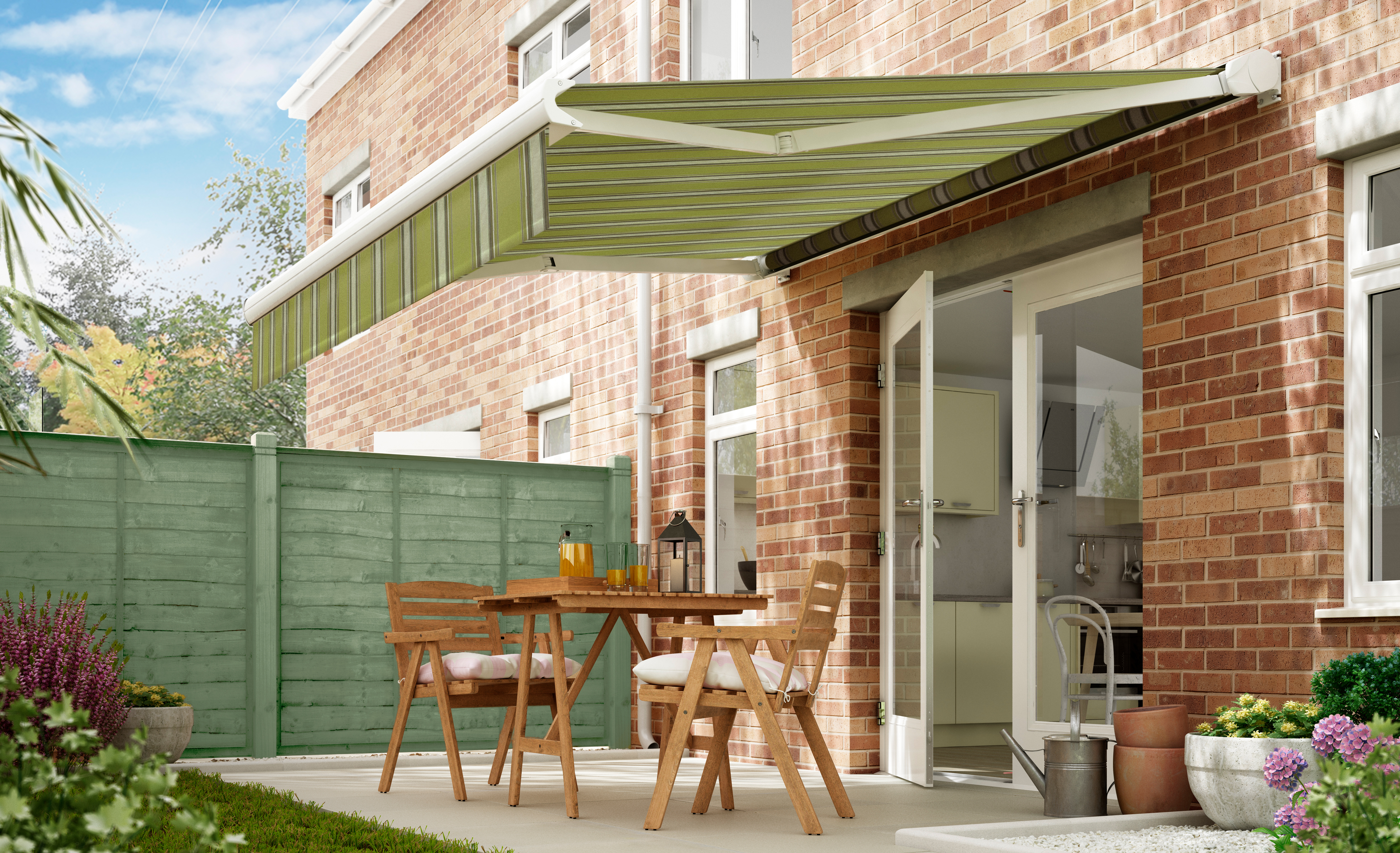 A green retractable awning shades a patio with wooden furniture outside a brick house, near a green fence and garden plants.