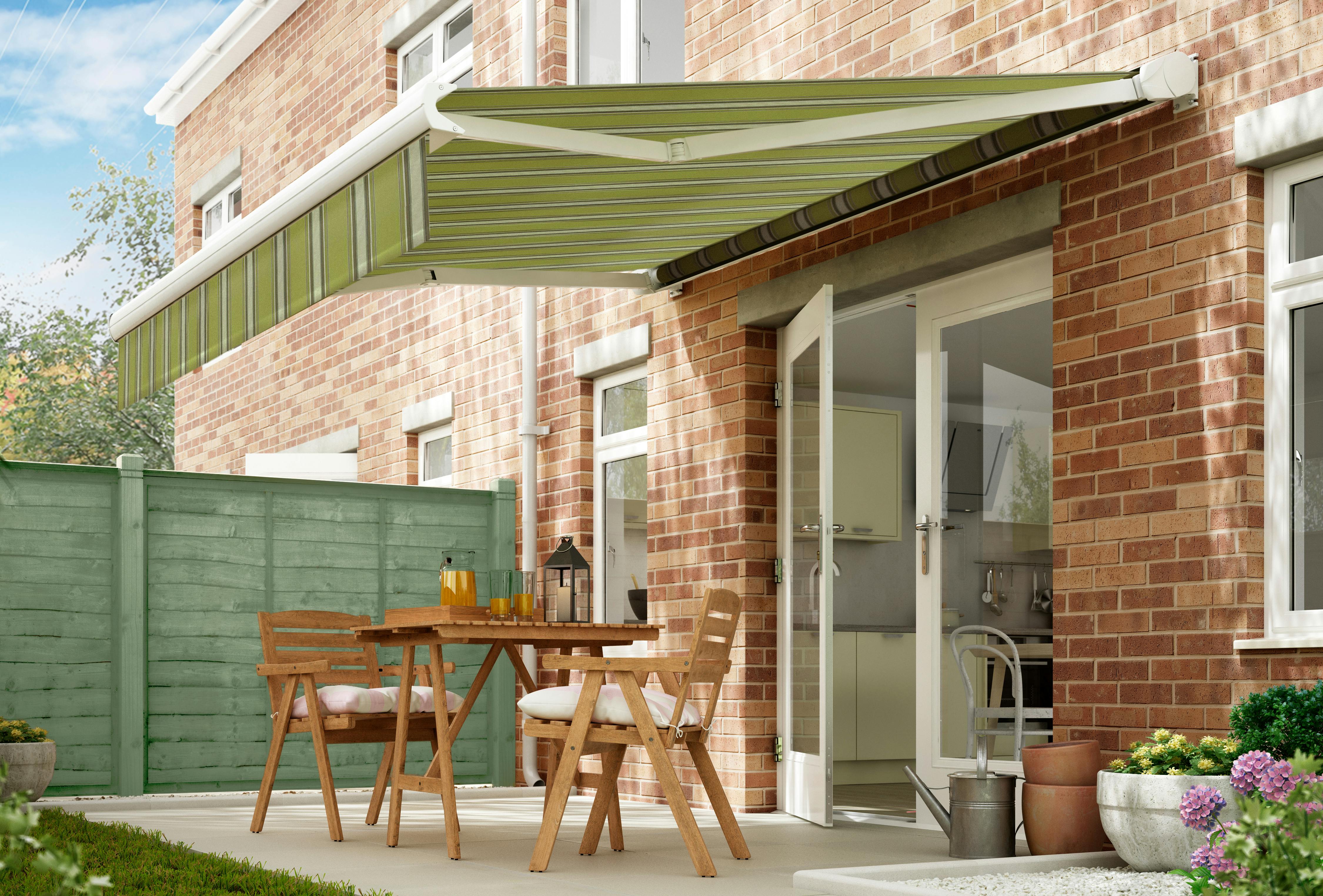 A green retractable awning shades a patio with wooden furniture outside a brick house, near a green fence and garden plants.