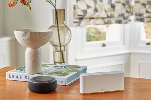 A white Somfy device rest on a wooden table in a room. Nearby is a ceramic bowl on books labeled "LEAF SUPPLY" and a glass vase with flowers by a window.