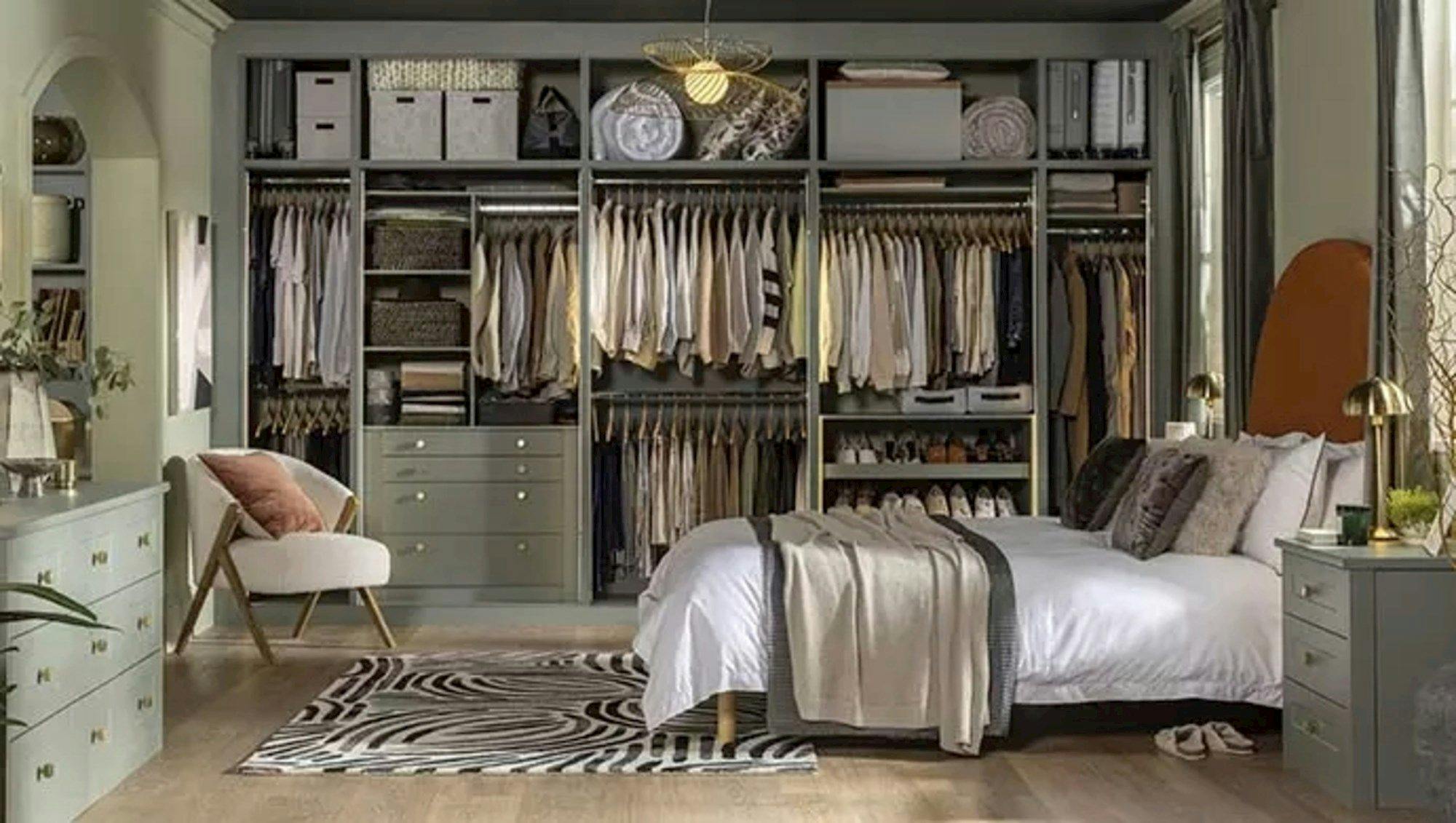 Ivory bedroom with wall to wall fitted units in mid grey showing racks of clothing, and storage baskets, bed with white bedding and ecru and grey throws, matching bedside table and chest of drawers on opposite wall, wooden floor with cream rug with dark grey linear pattern.