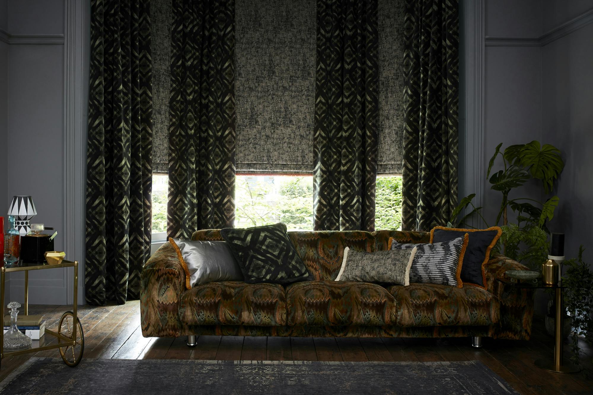 A dark, eclectic living room with patterned curtains, a velvet patterned sofa, assorted cushions, wood flooring, and lush green plants.