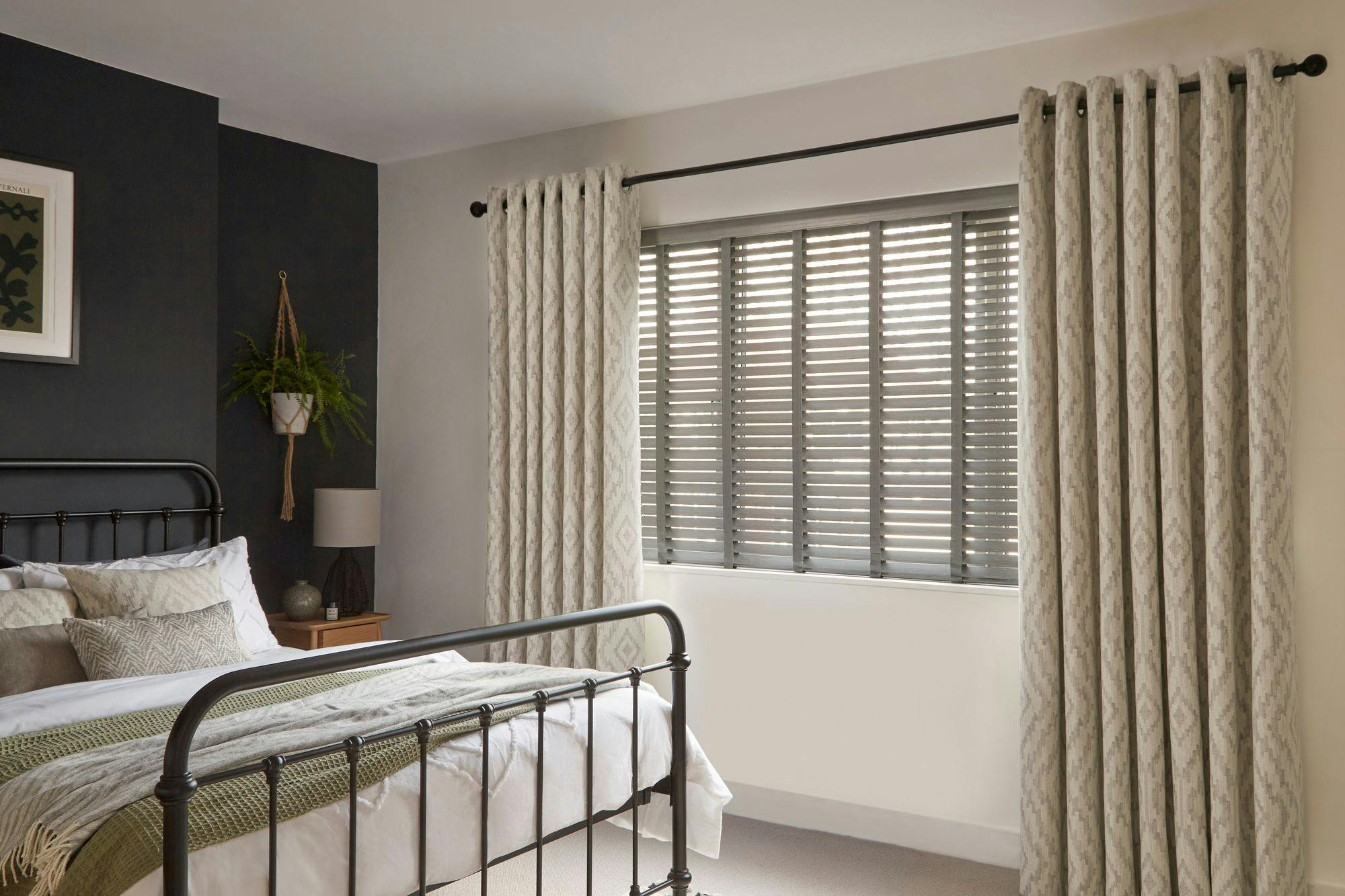A modern bedroom with charcoal wood Venetian blinds, light patterned curtains, a black metal bedframe, neutral bedding, and a potted hanging plant.