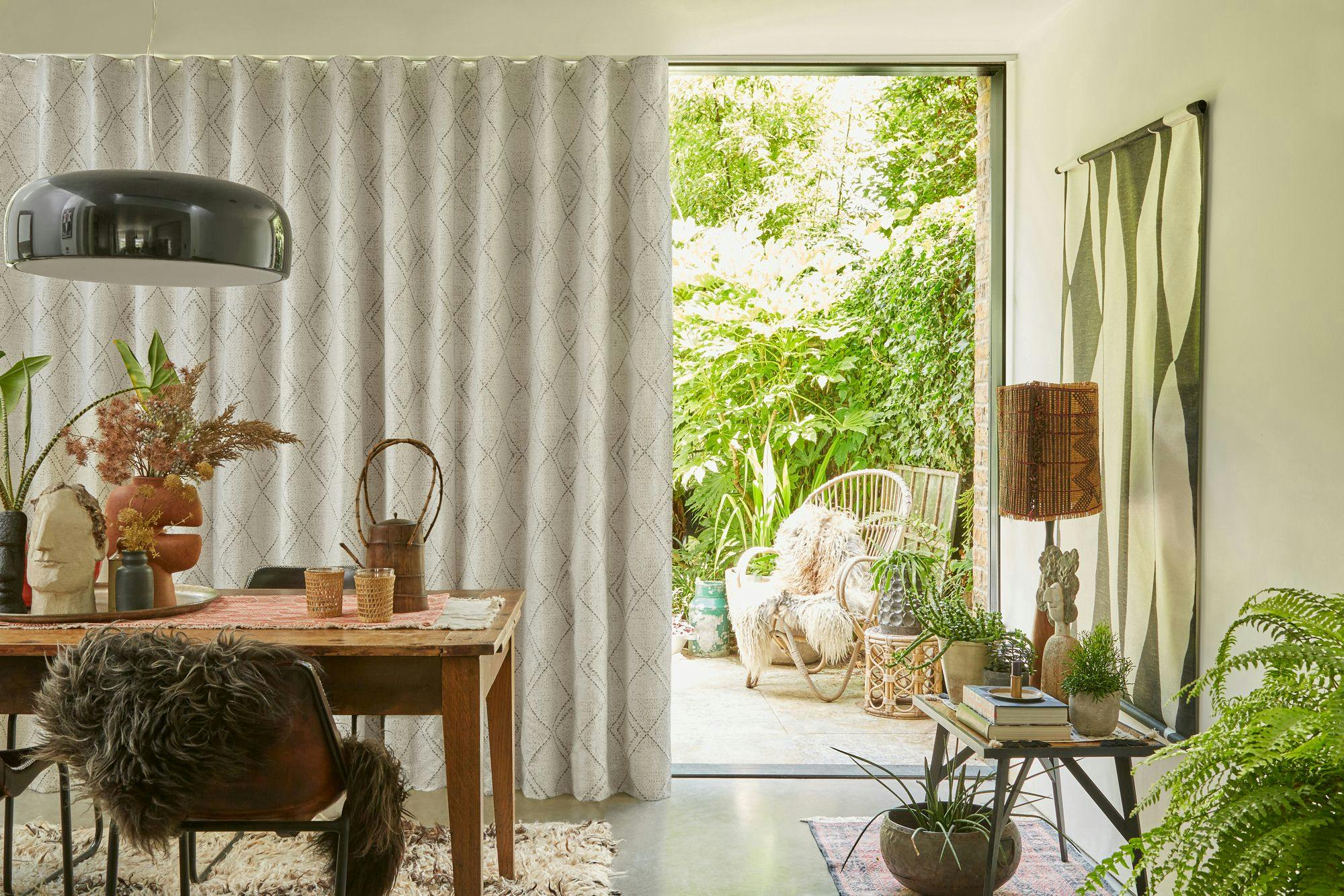 A bohemian-style dining space with light wave-patterned curtains, natural wood furniture, eclectic decor, and an open view to a lush garden patio.
