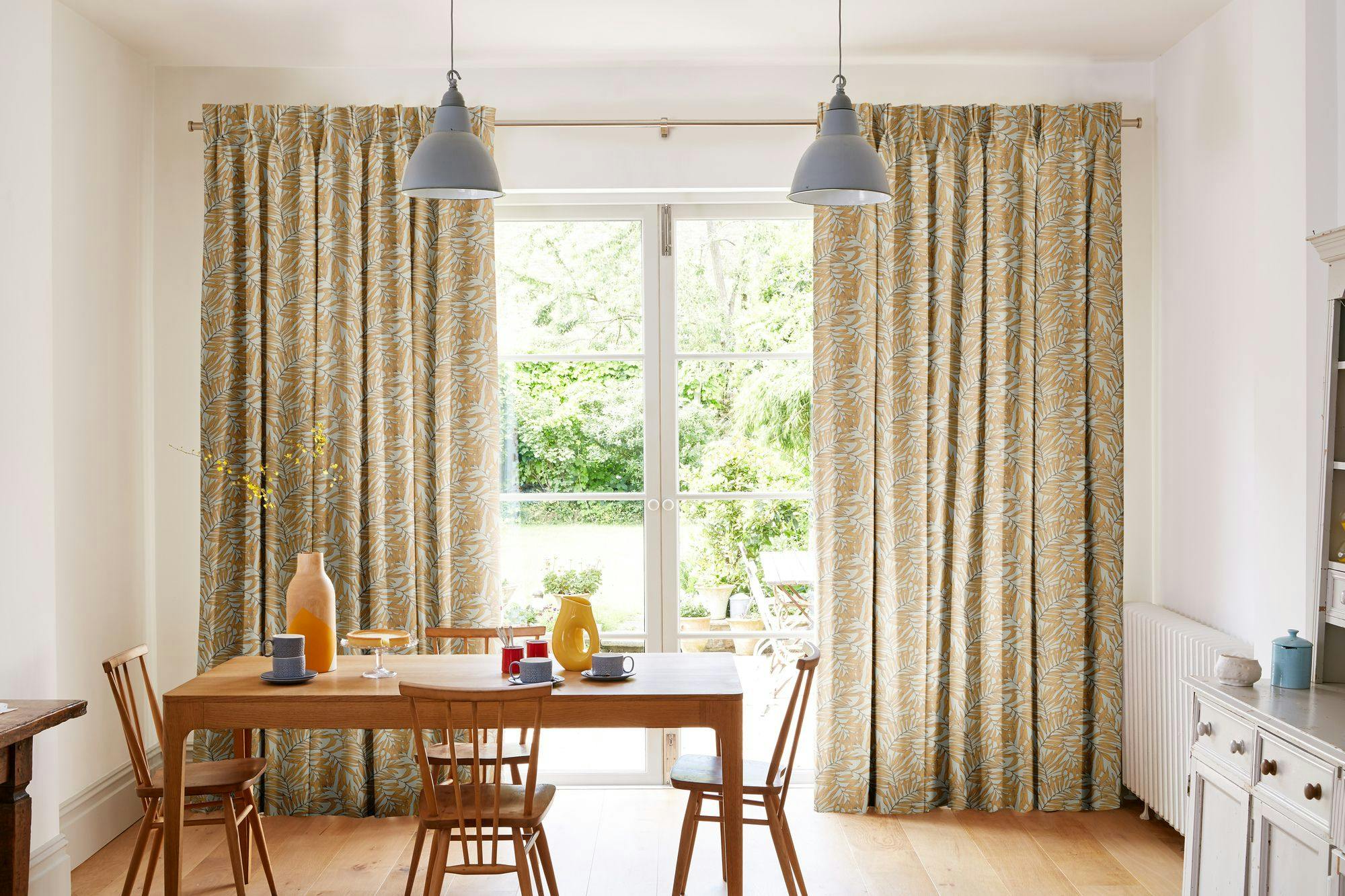 Bright dining room with yellow patterned curtains, wooden furniture, pendant lights, and large glass doors opening to a lush green garden.