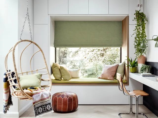 A Window seat with green cushions overlooks plants through a large window with a green Roman blinds. Nearby, a swing chair and a desk with plants and books complete the cozy room.