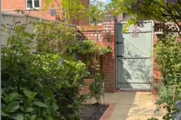 View from under a pink netted and fringed parasol down a garden path with tall shrubbery looking at a sage green garden gate in a red brick wall.