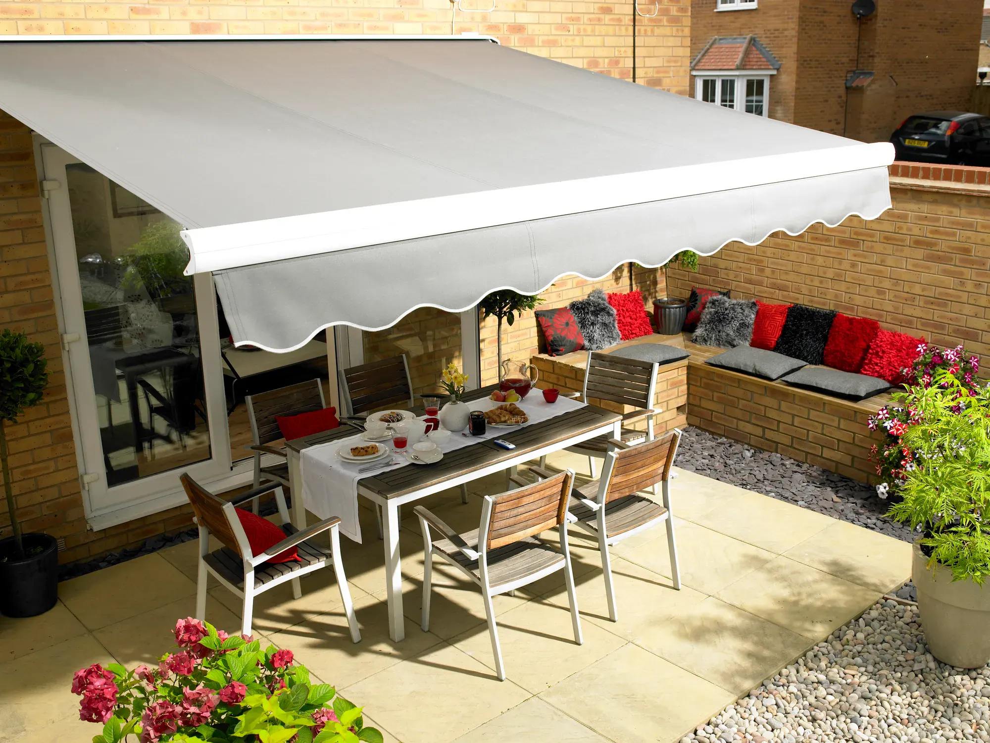 Grey retractable awning extended over a tiled patio, shading an outdoor dining set beside a brick wall and built-in barbecue area.