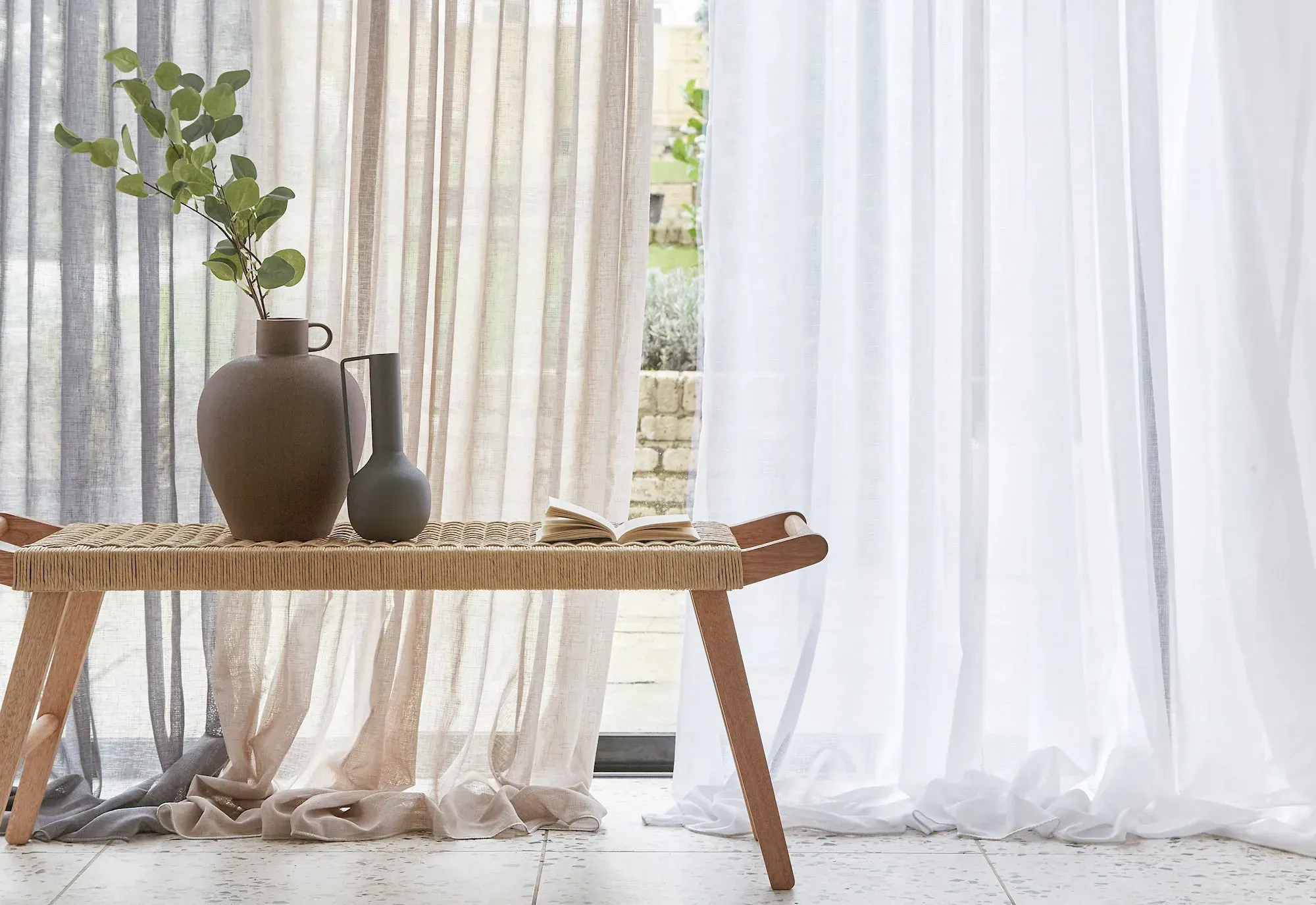 Soft sheer curtains in neutral tones framing a window, with a woven bench in front decorated with simple ceramic vases and greenery.