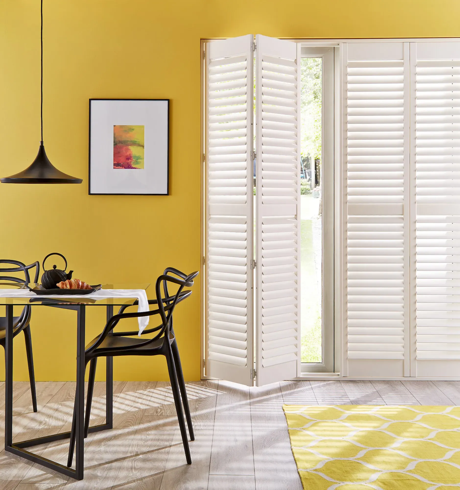 A bold, sunlit dining room with mustard yellow walls, white full-length shutters on patio doors and sculptural black dining chairs.
