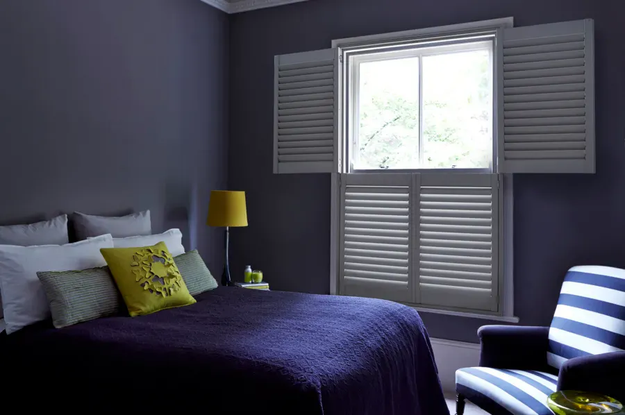 A bold, moody bedroom with deep violet walls and a grey-painted sash window shutter, offset by acid yellow cushions and a navy and white striped armchair.