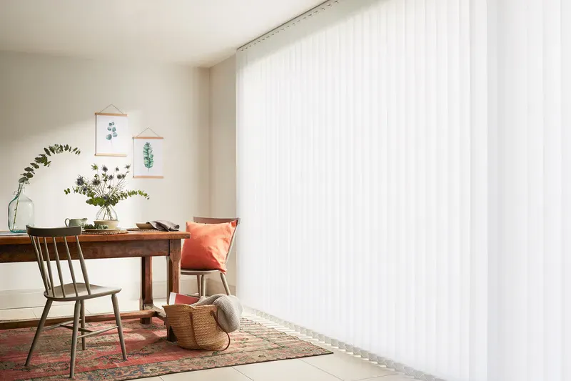 Large window with white Vertical blinds softly filtering daylight into a cosy dining nook with wooden table, chairs and patterned rug.