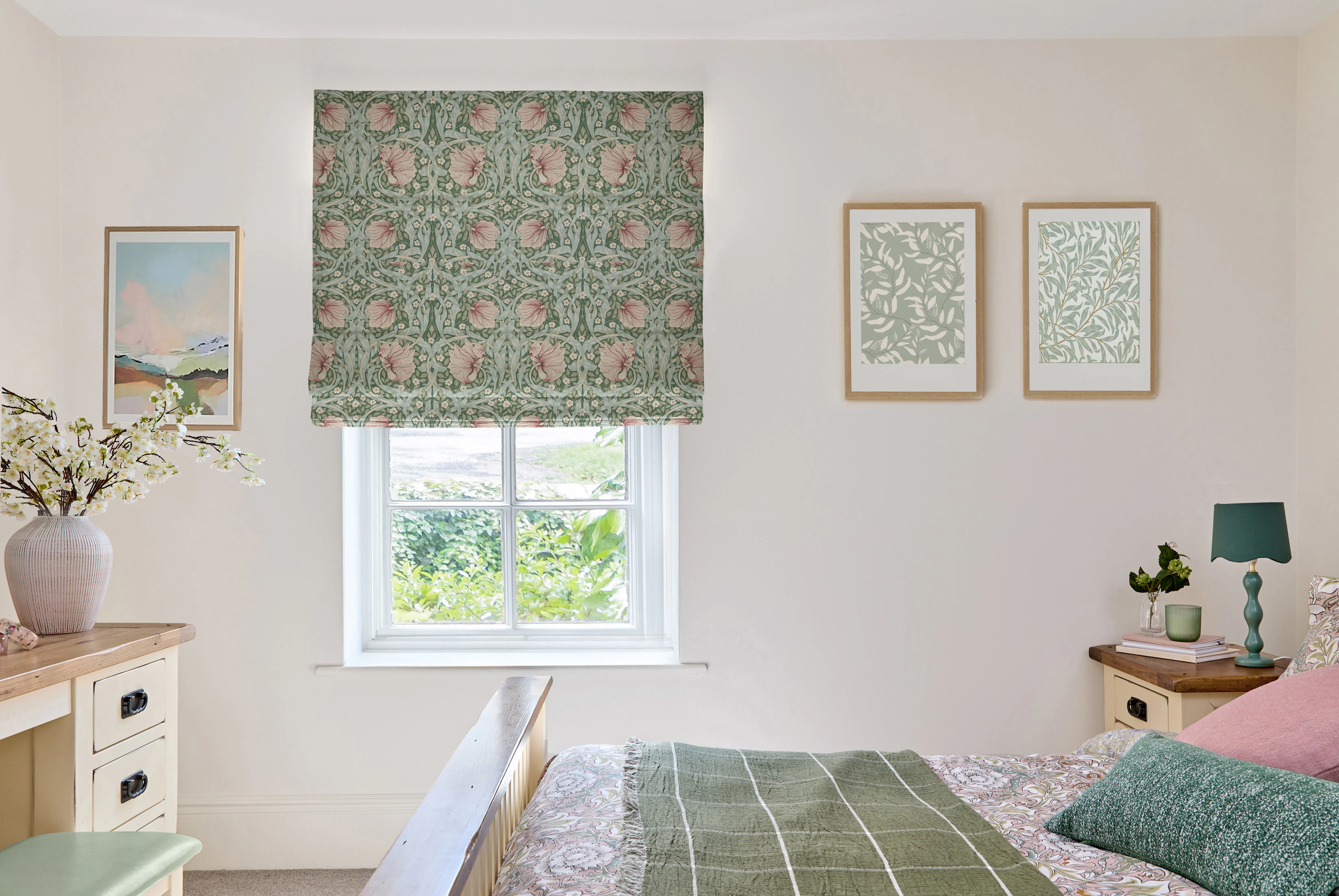 Bedroom window with a floral Roman blind, above a neatly made bed, with framed artwork and soft neutral walls.