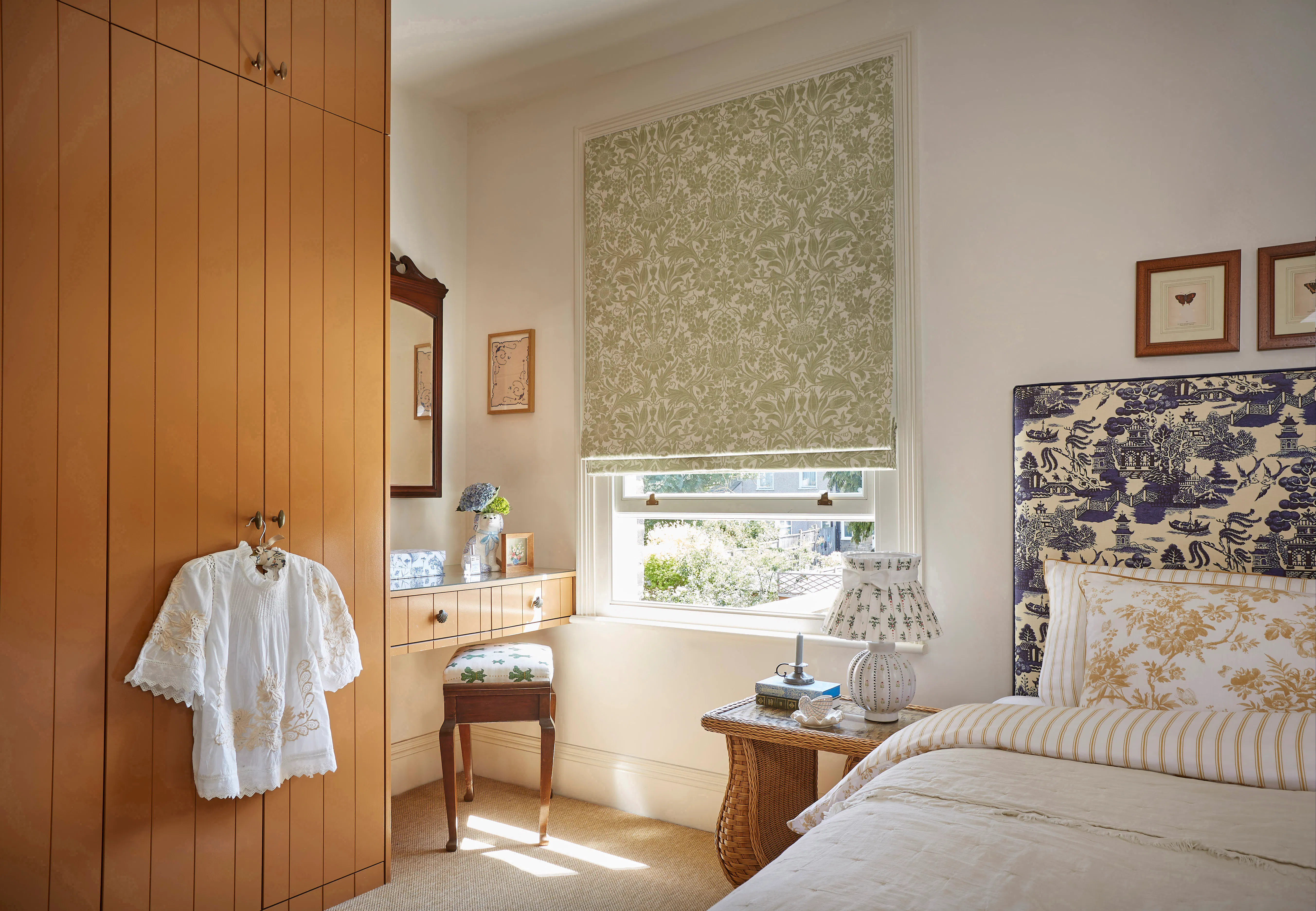 Green patterned Roman blind over a bright bedroom window, with wicker bedside table, upholstered bed and warm timber wardrobe.