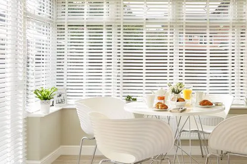 Bright bay window dressed with white blinds, filtering daylight into a modern breakfast area with a small round table, chairs and morning pastries.