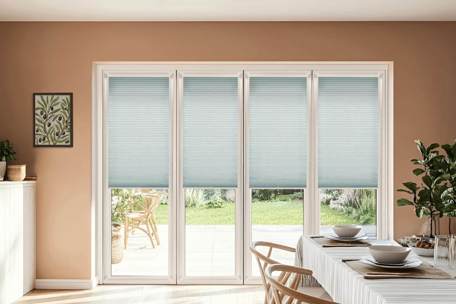 Magnolia dining room with white-framed bifold doors, closed, dressed in pale blue duette blinds lowered halfway, table with white tablecloth with navy blue pinstripe bands, wooden chairs, white dinner service, sunlight on wooden floor.