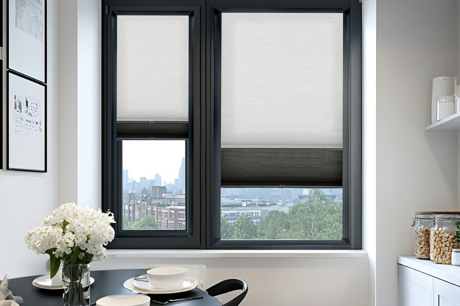 Close in on white kitchen end, with two adjacent windows in black frame dressed in transition blinds, white upper, lowered to middle, black lower raised to middle on left, partially raised on right, white base units and shelving on right, black round table and two chairs on left, white crockery and flowers on table, black and white geometric patterned rug on floor.