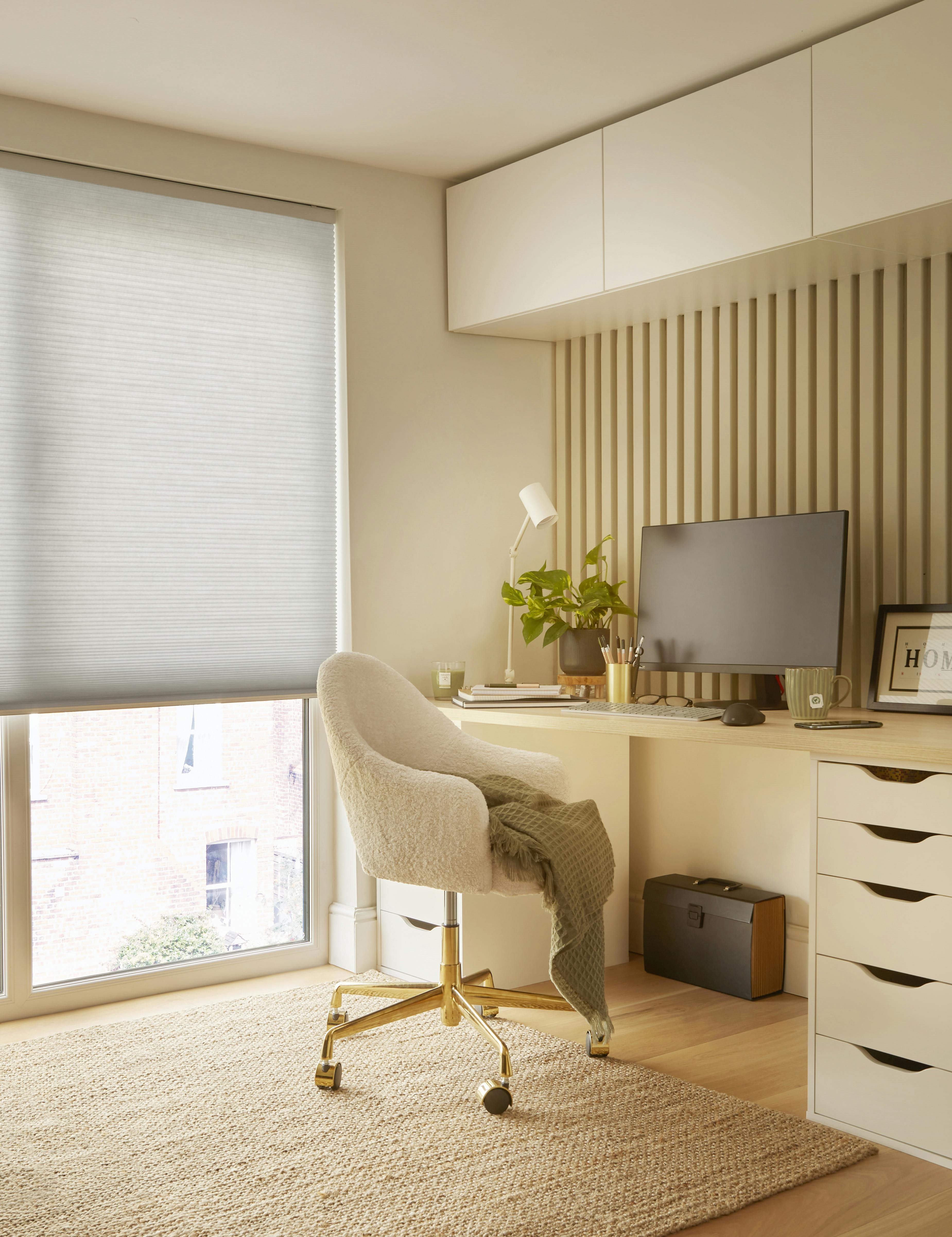 Ivory home office with floor to ceiling window dressed in pale grey duette blind, lowered halfway, ivory desk with drawers, high units above, matching chair, jute rug on wooden floor, monitor, small plant and desk lamp.