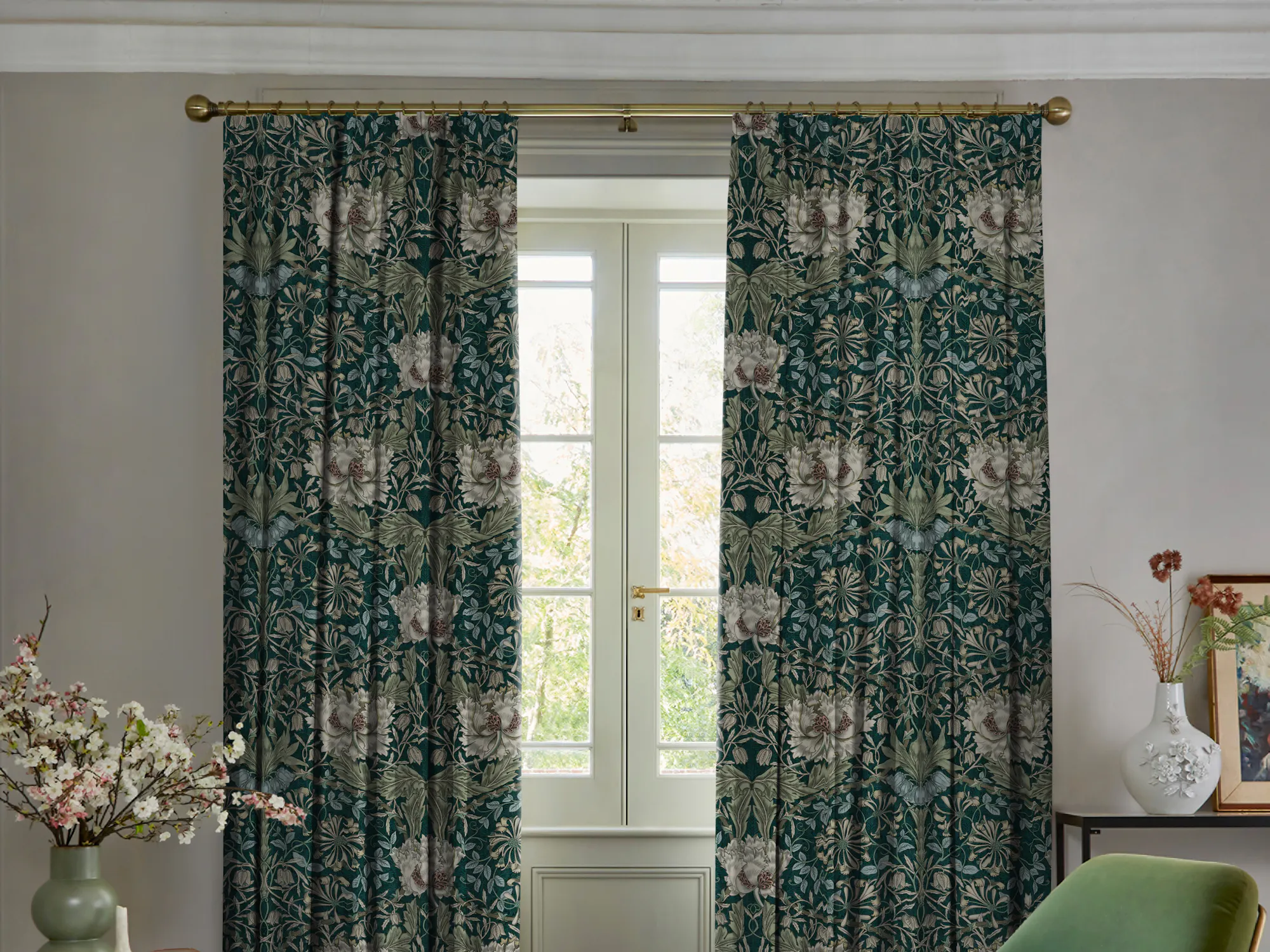 Living room in pale mink with curtains in dark green with cream floral pattern, partly open, polished concrete floor, green velvet occasional chair, with pale mink throw over arm, round table with green vase with foliage.