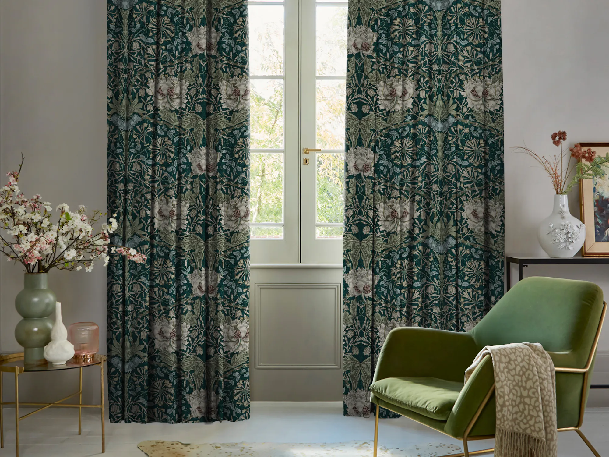 Living room in pale mink with curtains in dark green with cream floral pattern, partly open, polished concrete floor, green velvet occasional chair, with pale mink throw over arm, round table with green vase with foliage.