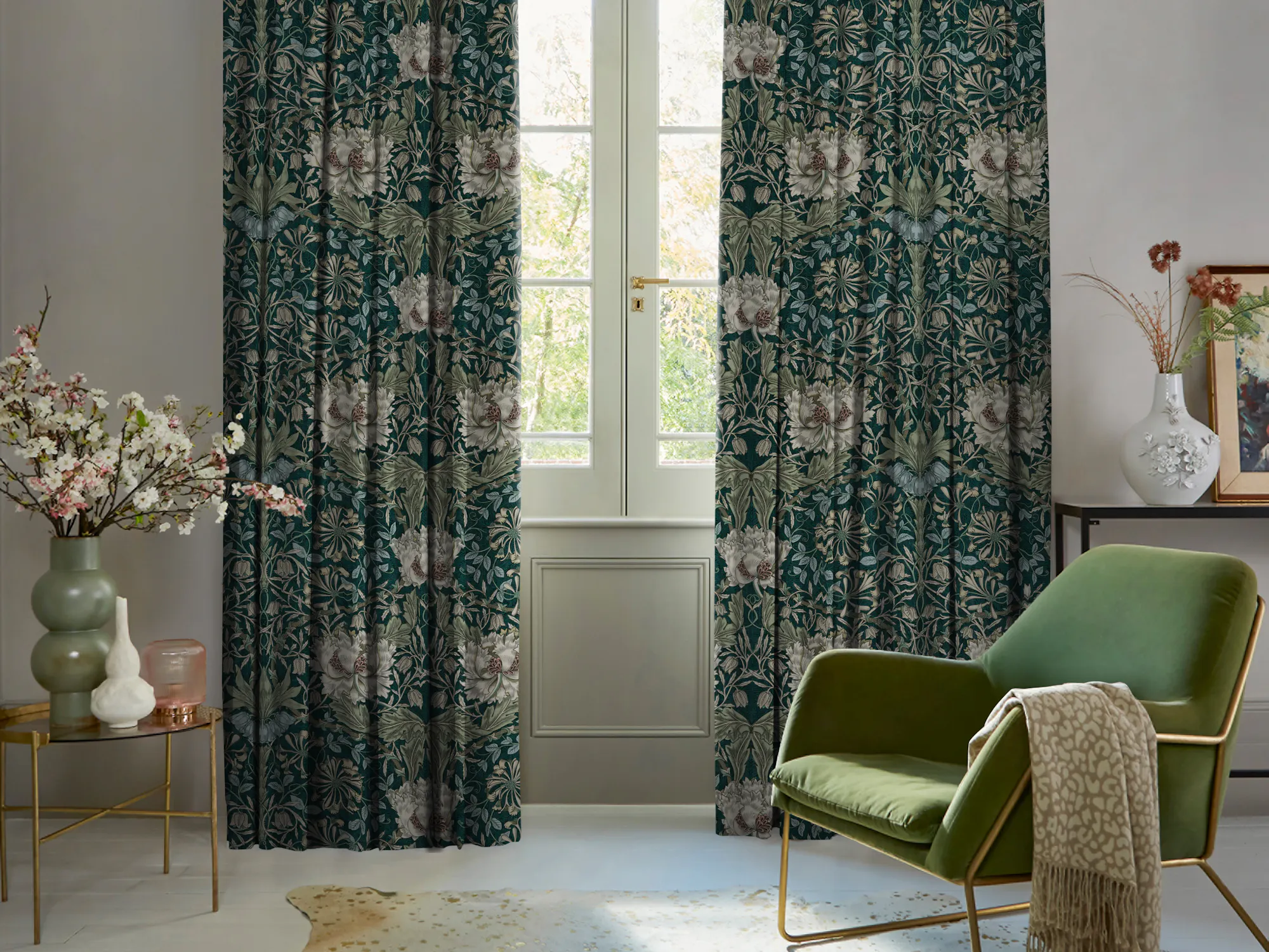 Living room in pale mink with curtains in dark green with cream floral pattern, partly open, polished concrete floor, green velvet occasional chair, with pale mink throw over arm, round table with green vase with foliage.