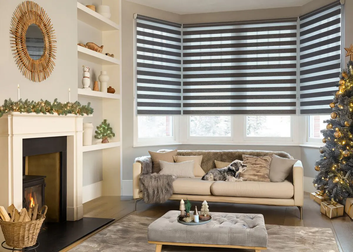 A neutral bay-window living room with a black-and-white striped day-and-night blind, a cream sofa, a sunburst mirror, and a gold-lit Christmas tree in the corner.