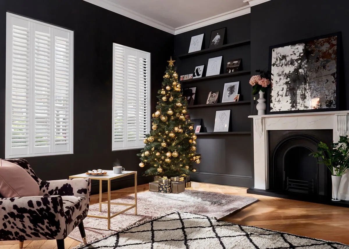 A dark living room with white plantation shutters, a gold-decorated Christmas tree beside a marble fireplace, and a patterned armchair on a soft pink rug.
