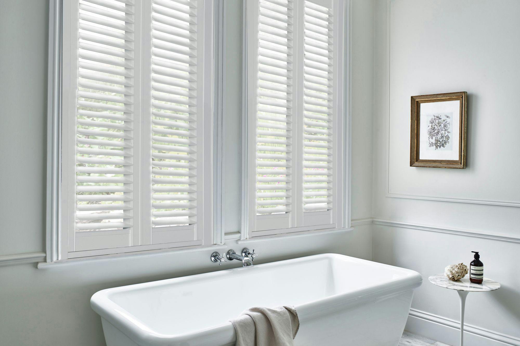 Bright white bathroom with vinyl shutters, freestanding bathtub, wall art, and a small marble table with soap and lotion.