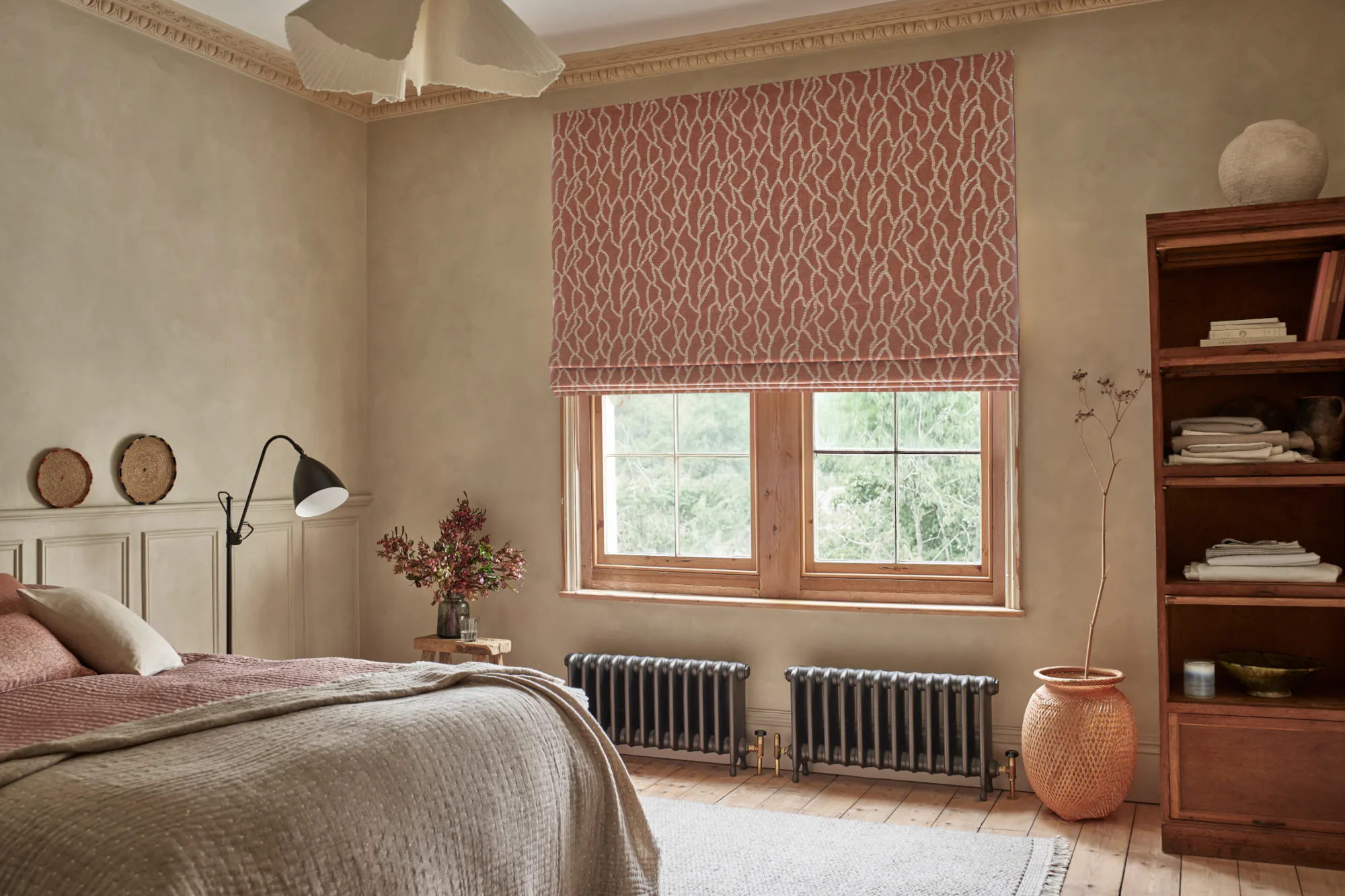 Bedroom with taupe plaster walls, window with roman blind in cream with rust pattern, tape throw on bed, vase on wooden floor.