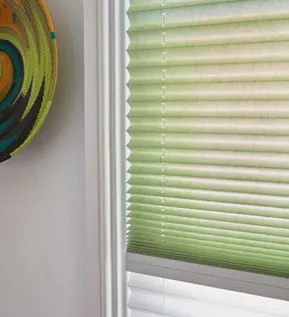 Close-up of green pleated blind on a white-framed window with a colourful circular wall decoration nearby.