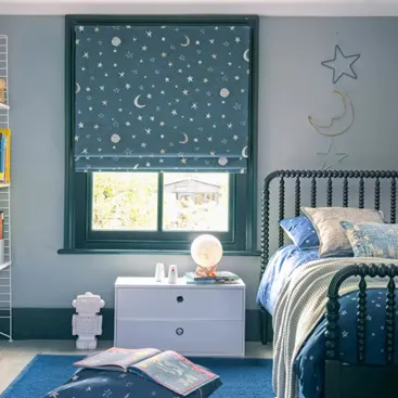 Children’s bedroom with blue Roman blind featuring stars and moons, matching bedding, and a white bedside table.