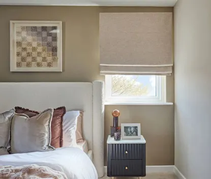 Bedroom with beige Roman blind on window, neutral walls, and a bed with layered cushions beside a small black bedside table.
