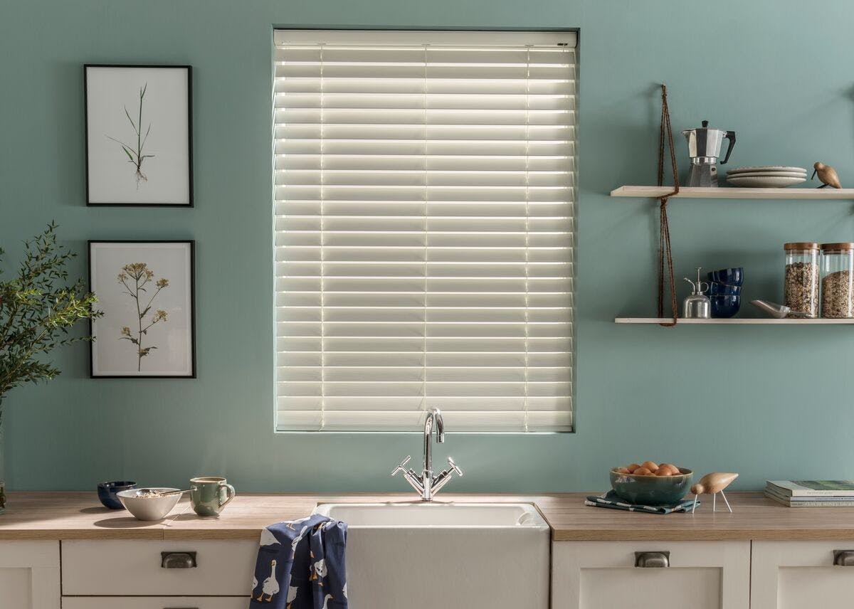 Kitchen with a window featuring cream venetian blinds above a sink, with shelves, plants and tableware on light wooden countertops.