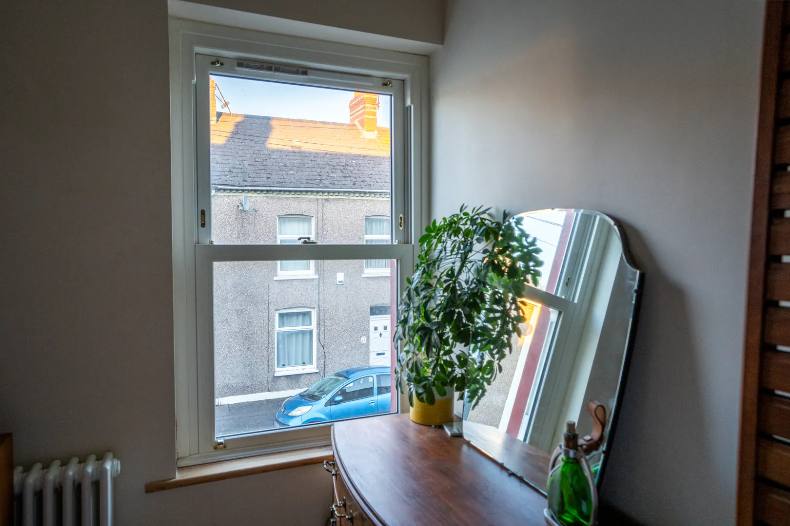 View through a window onto a terraced house and parked blue car, with a plant and mirror on a wooden dresser in the foreground.