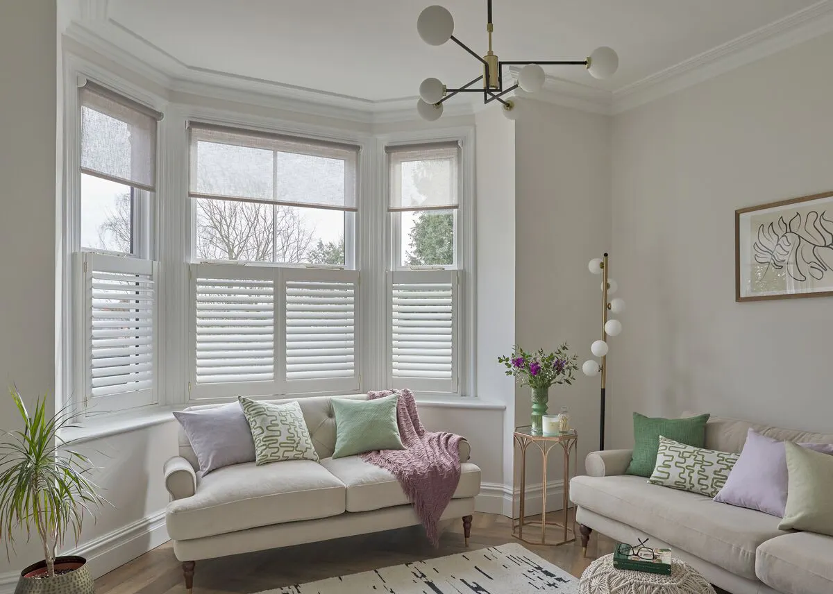 Bright living room with bay windows, white shutters, light sofas with pastel cushions, plants, and modern décor